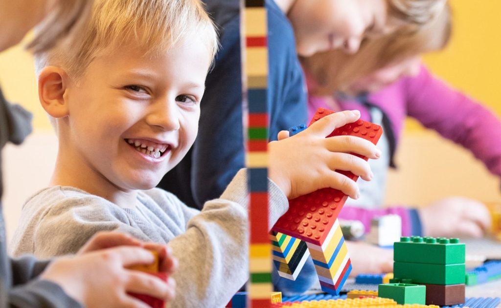 Ein lächelndes Kind in einem grauen Hemd spielt mit bunten Bauklötzen an einem Tisch. Im Hintergrund bauen auch andere Kinder. Die Szene strahlt eine fröhliche und kreative Atmosphäre aus.