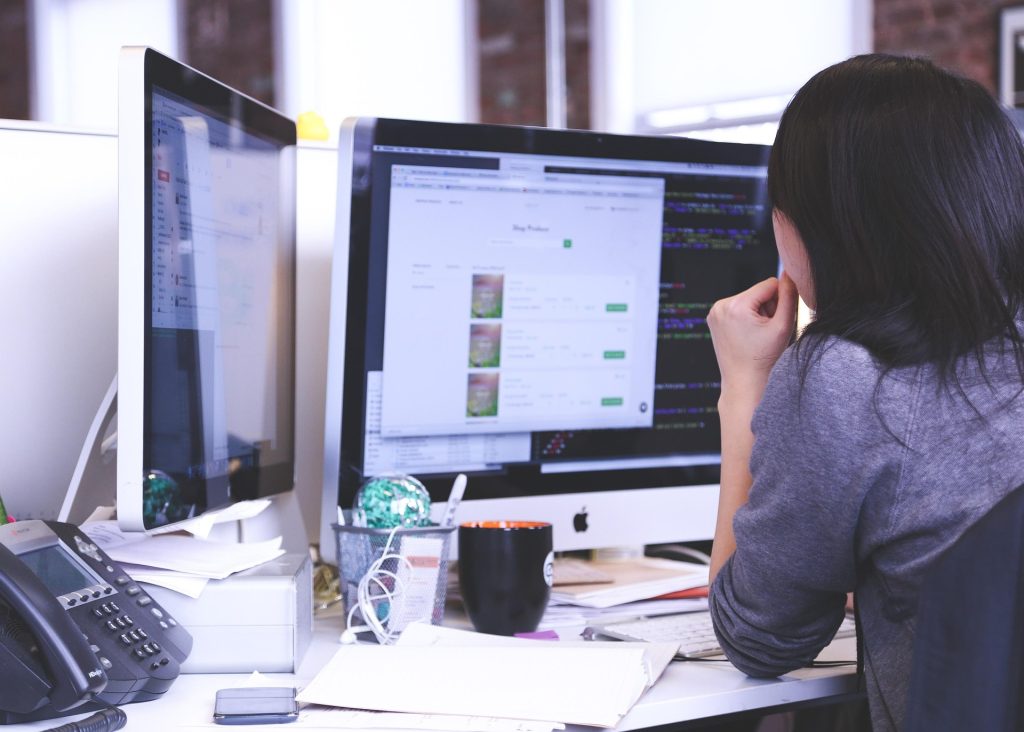 Eine Person sitzt an einem Schreibtisch und blickt auf zwei Computerbildschirme. Ein Bildschirm zeigt eine Programmieroberfläche, der andere eine Online-Shop-Seite. Auf dem Schreibtisch liegen Telefon, Notizblock, Stift und Tasse. Das Büro ist modern und minimalistisch gestaltet.