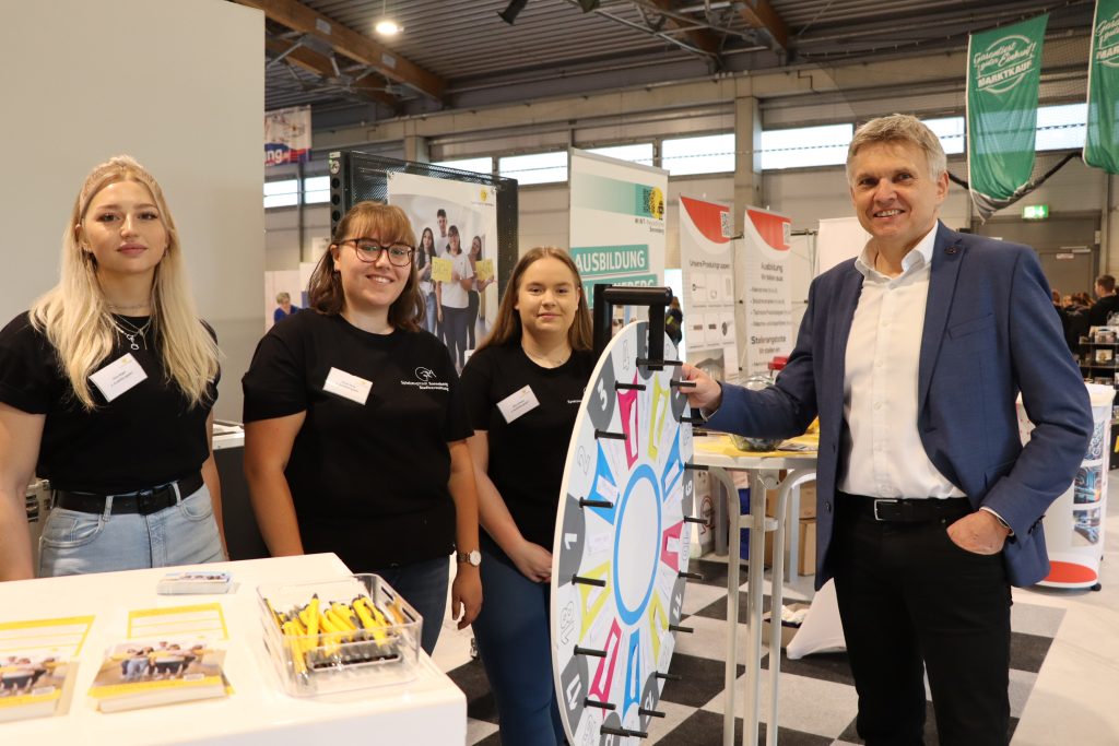 Auf dem Bild sehen Sie 3 junge Frauen auf der FAMOS mit ihrem Messestand. Am MINT-Rad steht der Bürgermeister der Stadt Sonneberg. Im Hintergrund sehen Sie die Eishalle mit anderen Werbebannern von Ausstellern.