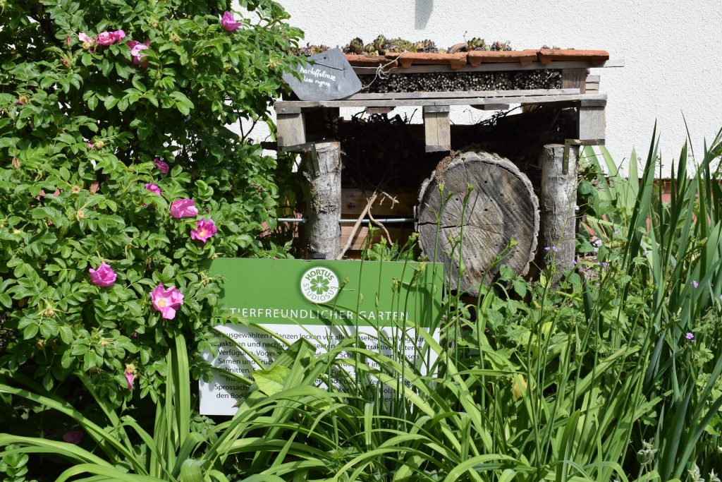Sie sehen einen tierfreundlichen Garten mit zahlreichen Blüh- und Grünpflanzen. In der Mitte steht versteckt eine kleine Informationstafel und im Hintergrund ein Insektenhotel aus verschiedenen Naturstoffen.