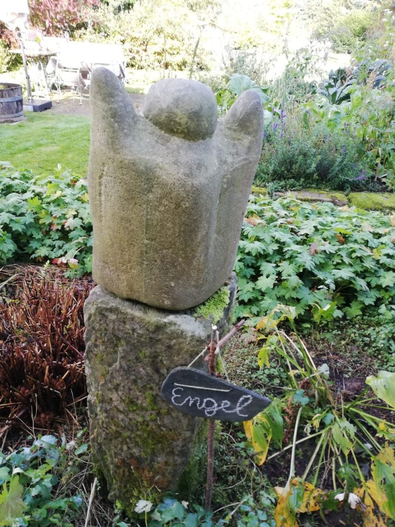 In der Mitte steht eine Skultptur aus Stein in mitten eines blühendes Gartens.