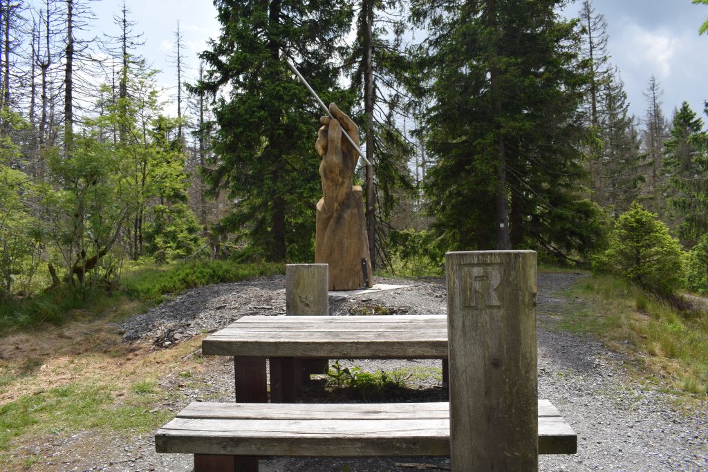 Im Vordergrund befindet sich eine Holzsitzgruppe auf geschotterter Fläche. Im Hintergrund steht vor dem Wald eine Holzskulptur. Diese Holzslulptur symbolisiert eine Hand, die einen SChreibgriffel hält.