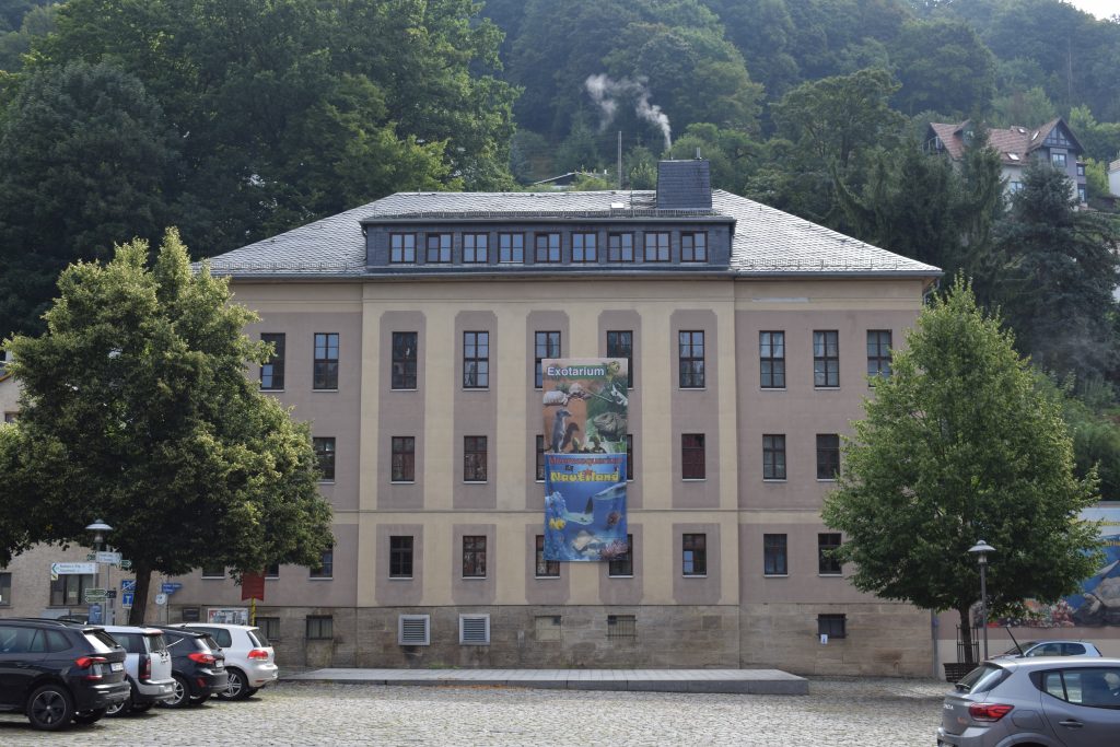 Vor einem grün bewachsenen Hang steht das beige dreistöckige Gebäude des Nautiland. Davor parken Autos links und rechts auf einem Parkplatz.