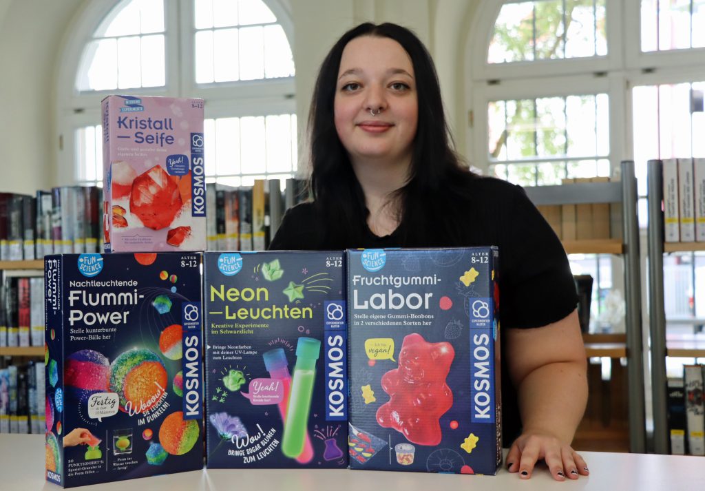 Experimentboxen stehen auf einem Tisch. Im Hintergrund steht eine junge Frau mit schwarzen Haaren vor dem Fenster.