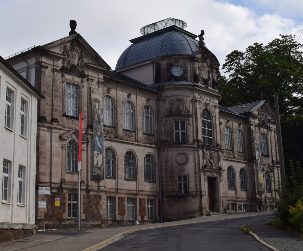 Dreistöckiges Sandsteingebäude mit Kuppel auf dem Dach. Im HIntergrund wolkenbehangender Himmel und davor ein Fußweg und Straße.