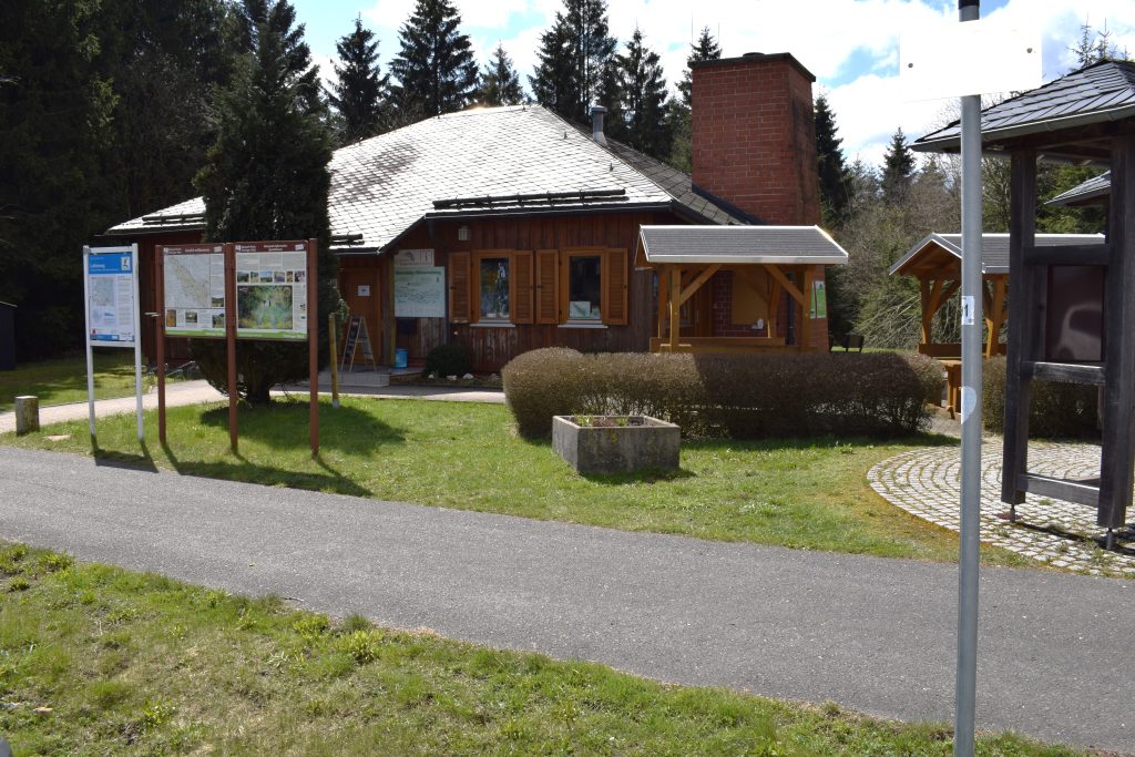 Ein Holzgebäude steht auf einer grünen Wiese und besitzt ein Schieferdach. Davor stehen Informationstafel und Sitzmöglichkeiten. Im Hintergrund blauer Himmel mit Wolken und eine Baumreihe.