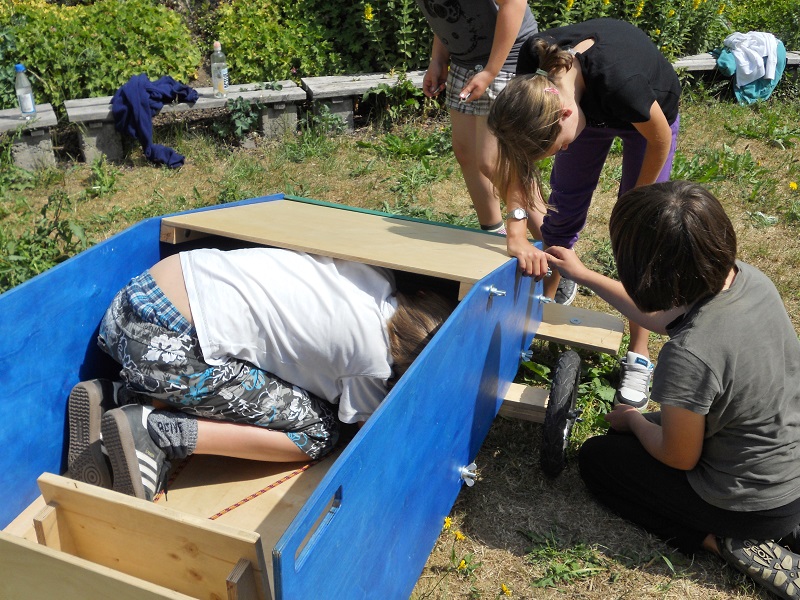 Drei Personen bauen eine Seifenkiste aus Holz auf einer Wiese.